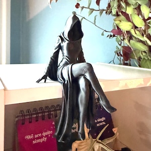 Pode incluir: Uma estatueta preta fosca de uma figura encapuzada com um manto longo, sentada com as pernas cruzadas. A figura tem um capuz profundo e sombreado e mangas largas, com um acabamento liso e sólido. O design retrata um personagem sentado.