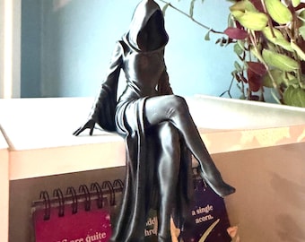 May include: A matte black figurine of a hooded figure in a long robe, seated with legs crossed. The figure has a deep, shadowed hood and flowing sleeves. Below the figure, a small sign features the text: We are quite simply a single acorn.