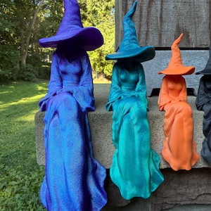 May include: A set of four witch figurines in varying sizes, each wearing a wide-brimmed pointed hat and a long flowing gown. The figures are colored in vibrant purple, teal, orange, and black, featuring a textured surface finish throughout.