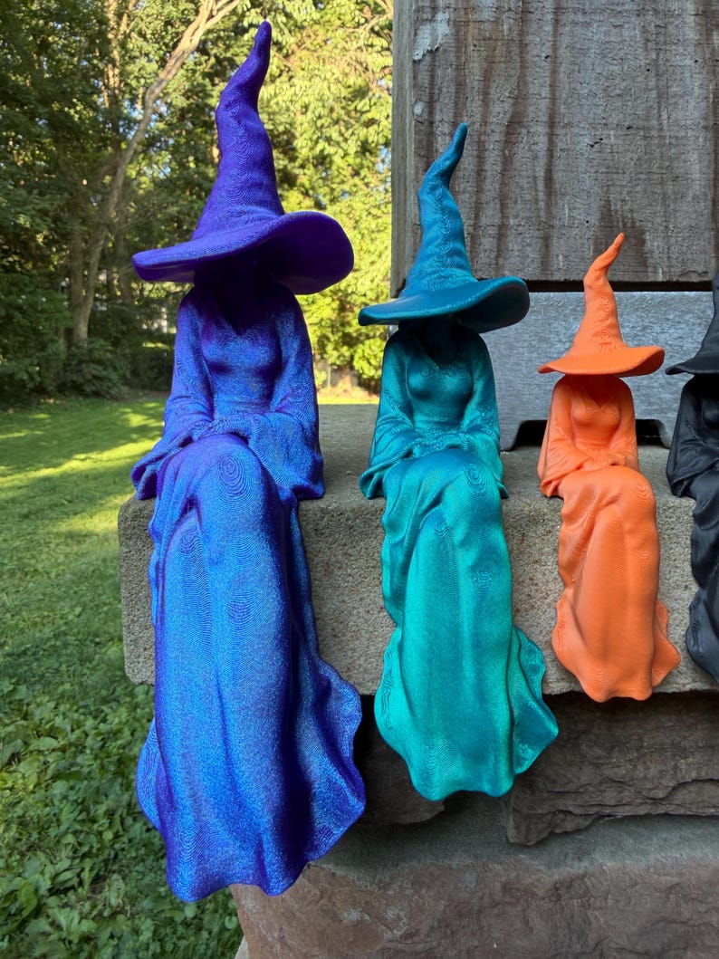 May include: A set of four witch figurines in varying sizes, each wearing a wide-brimmed pointed hat and a long flowing gown. The figures are colored in vibrant purple, teal, orange, and black, featuring a textured surface finish throughout.