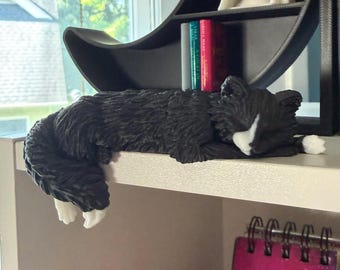 May include: A black and white cat figurine in a sleeping position, featuring textured fur details, a white patch on the face, and white paws. The cat is positioned on the edge of a shelf, with its tail hanging down over the side of the surface.
