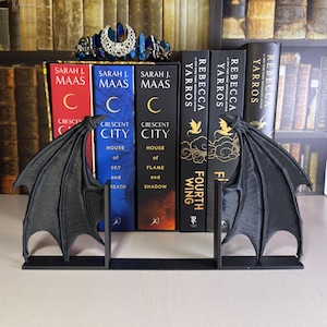May include: Black, wing-shaped bookends stand before a row of books. The bookends have a detailed, textured design. The books include titles by Sarah J. Maas and Rebecca Yarros. A blue and silver crown sits on the shelf above.