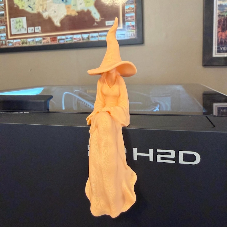 May include: A peach-colored figurine of a seated figure wearing a long, flowing gown and a tall, pointed hat. The figure is positioned with legs crossed at the knee, featuring a smooth, matte texture and a solid, monochromatic appearance.