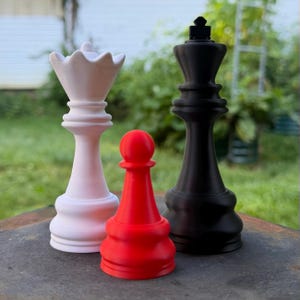 May include: Three large chess pieces standing together: a white queen on the left, a red pawn in the center, and a black king on the right. Each piece features a smooth, matte finish and a classic design with a wide base and distinct top shapes.