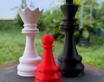 May include: Three large chess pieces standing together: a white queen on the left, a red pawn in the center, and a black king on the right. Each piece features a smooth, matte finish and a classic design with a wide base and distinct top shapes.