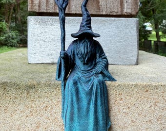 May include: A dark blue and black 3D printed figurine of a wizard wearing a pointed hat and long robes, holding a tall staff with a textured top. The figure features a seated posture with a long beard and detailed fabric folds in the clothing.