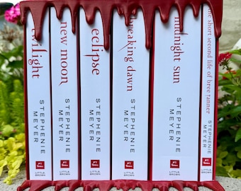 May include: A set of six Twilight series books by Stephenie Meyer, including Twilight, New Moon, Eclipse, Breaking Dawn, Midnight Sun, and The Short Second Life of Bree Tanner, held in a dark red bookend with a decorative dripping blood effect.