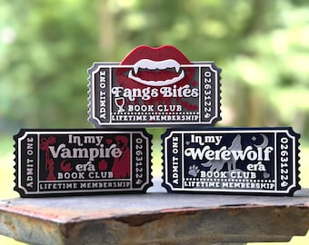 May include: Three rectangular ticket-shaped plaques featuring raised text and graphics. The top plaque shows red lips with fangs and reads Fangs Bites Book Club Lifetime Membership. The bottom left reads In my Vampire era Book Club Lifetime Membership. The bottom right reads In my Werewolf era Book Club Lifetime Membership.