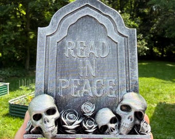 May include: A gray tombstone-shaped decorative object with a weathered finish featuring the embossed text READ IN PEACE. The base is adorned with three human skulls and several sculpted roses, all finished in a matching metallic gray tone.