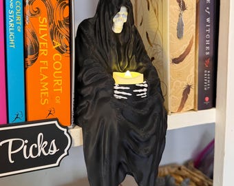 May include: A black Grim Reaper figurine with a skull face and a draped robe, holding a glowing candle. The figurine is perched on a shelf, with colorful book spines visible in the background. The word "Picks" is on a black and white sign.