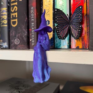 May include: A purple 3D printed figurine of a seated figure wearing a pointed hat and a long flowing robe, positioned next to a black and purple butterfly decoration. The figure has a smooth, iridescent finish and is displayed on a shelf.