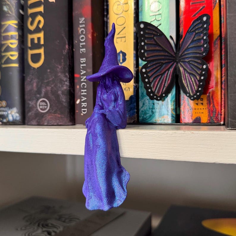 May include: A purple 3D printed figurine of a seated figure wearing a pointed hat and a long flowing robe, positioned next to a black and purple butterfly decoration. The figure has a smooth, iridescent finish and is displayed on a shelf.