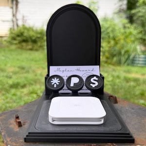 May include: A black arched display stand featuring three circular icons with white symbols for a star, the letter P, and a dollar sign. A white square payment card reader sits on the base in front of a business card labeled Meghan Howard.