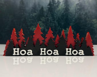 May include: A decorative tabletop display featuring a layered forest silhouette with red and black pine trees. The front black layer displays the white text "Hoa Hoa Hoa" in a bold, sans-serif font, creating a festive, multi-dimensional scene.