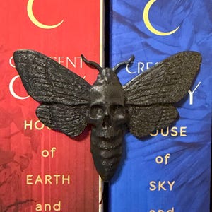 May include: A dark, textured moth ornament featuring a central skull design on its thorax, positioned against two books with red and blue covers. The books display the text Crescent City, House of Earth and Blood, and House of Sky and Breath.