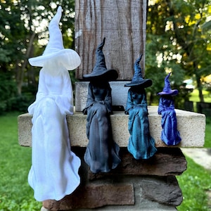 May include: Four witch figurines in descending order of size, each wearing a pointed hat and a long flowing gown. The first is white, the second is matte black, the third is a metallic teal, and the smallest fourth figurine is a vibrant royal blue.