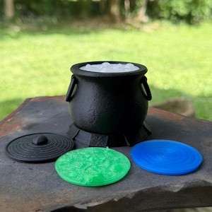 May include: A black cauldron with a textured surface and two side handles, containing a white bubbling liquid. Three circular flat lids in black, translucent green, and translucent blue are positioned in front of the cauldron base.