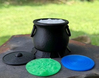 May include: A black cauldron with a textured surface and two side handles, containing a white bubbling liquid. Three circular flat lids in black, translucent green, and translucent blue are positioned in front of the cauldron base.