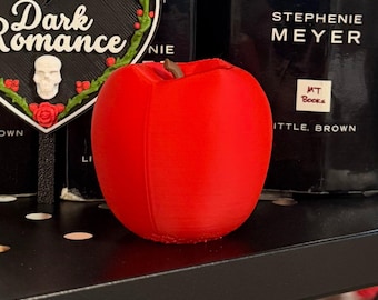 May include: A bright red, three-dimensional apple figurine with a small brown stem at the top. The apple has a smooth, matte finish and a rounded shape, positioned centrally in front of black book spines and a heart-shaped sign with text.