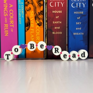 以下が含まれることがあります： 「To Be Read」の文字が白文字で書かれたビーズのブックチャーム。紫色のハート型のビーズが間に入っています。カラフルな本の列の前に配置されています。
