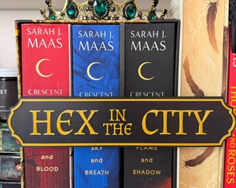 May include: A bookshelf display featuring books by Sarah J. Maas, with red, blue, and black covers, and a decorative sign that reads "HEX IN THE CITY." A gold crown with green jewels sits atop the books. The books are arranged on a shelf.