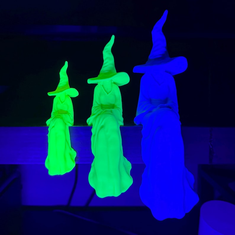 May include: Three witch figurines in varying sizes, each wearing a pointed hat and a long robe. The two smaller figures glow in a bright neon green, while the largest figure on the right glows in a vibrant blue, all set against a dark background.