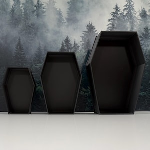 May include: Three matte black open containers shaped like coffins, arranged in a row from smallest to largest. Each container features a hexagonal silhouette with straight sides and a flat base, providing an open interior space for storage.
