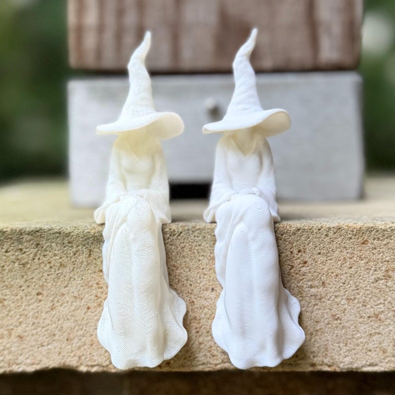 May include: Two white figurines of seated figures wearing pointed hats and long, flowing robes. The figures have a smooth, matte finish and are positioned side-by-side, each featuring a wide-brimmed hat and draped clothing with visible texture.