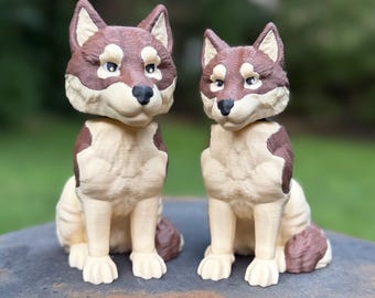 May include: Two sitting wolf figurines featuring a brown and cream color palette. Each wolf has pointed ears, a black nose, and detailed fur texture. The figurines are positioned side-by-side, displaying a consistent design with slight variations in size.