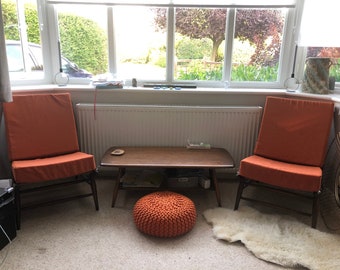 May include: Two orange chairs with wooden frames and a wooden coffee table with a woven orange ottoman in front of a window with a view of trees and a car.