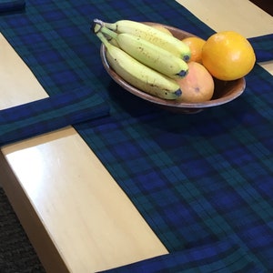 May include: A blue and green plaid table runner with a bowl of fruit on top. The bowl contains bananas, oranges, and a grapefruit. The runner is folded at the ends to create a decorative border.