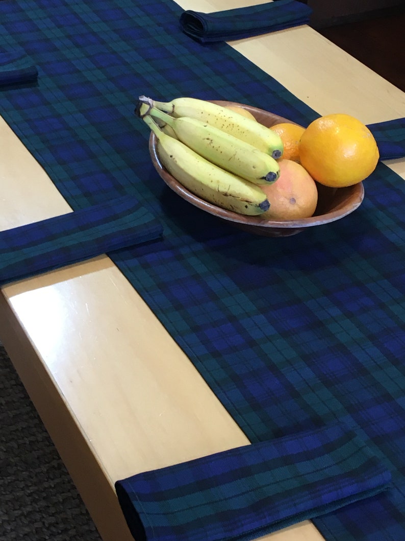 May include: A blue and green plaid table runner with a bowl of fruit on top. The bowl contains bananas, oranges, and a grapefruit. The runner is folded at the ends to create a decorative border.