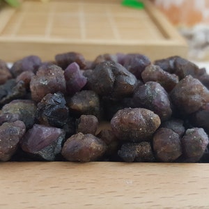 Op de afbeelding: Een verzameling ruwe, ongeslepen edelstenen in bruine en paarse tinten, rustend op een licht houten dienblad. De stenen variëren in grootte en vorm, met een natuurlijke, aardse uitstraling. De achtergrond bevat een houten dienblad en andere natuurlijke elementen.