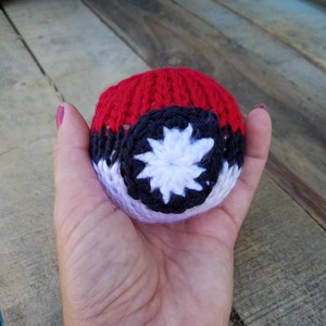 Può includere: Una palla fatta a mano, all'uncinetto, con una parte superiore rossa, una sezione centrale nera e bianca e un disegno a stella bianca. La palla è tenuta in una mano, mostrando i dettagli della lavorazione e i colori vivaci. L'oggetto è probabilmente un giocattolo o un elemento decorativo.