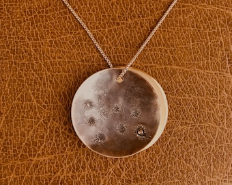 May include: A silver pendant necklace with a round, textured disc featuring a design of small, raised circles. The necklace is on a brown leather background.