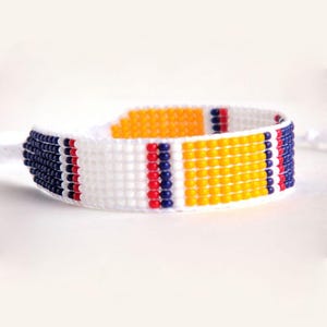 Op de afbeelding: Een geweven kralenarmband met een witte basis, versierd met strepen van marineblauwe, rode en gele kralen. De armband is ongeveer 2 cm breed. Een handgemaakt accessoire.