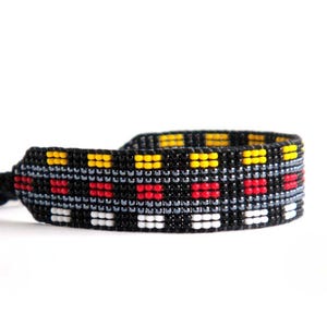 Op de afbeelding: Een zwart geweven armband met een herhalend patroon van rode, gele, witte en grijze vierkanten. De armband is gemaakt van kleine kralen en heeft een geknoopte sluiting.