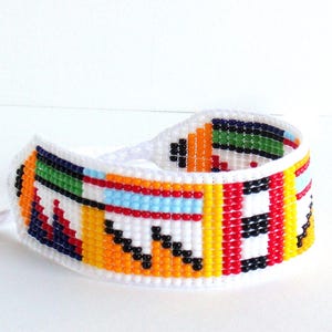 Op de afbeelding: Een kleurrijke kralenarmband met een witte basis. De armband heeft een geometrisch patroon met oranje, gele, rode, groene, blauwe en zwarte kralen. De armband is ongeveer 3,8 cm breed.