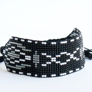 Op de afbeelding: Zwart-witte kralenarmband met een geometrisch patroon. De armband is gemaakt van kleine kralen en heeft een geweven ontwerp. Het heeft aan elk uiteinde een zwarte kwast.