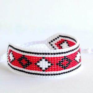 Op de afbeelding: Een handgemaakte armband met een geweven ontwerp. De armband heeft een rood, zwart en wit patroon. De hoofdgedeelte van de armband is rood met zwarte en witte geometrische vormen. De armband heeft een wit koord en kwast.