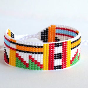Op de afbeelding: Een kleurrijke kralenarmband met een witte basis. De armband heeft een geometrisch patroon met rode, gele, groene, zwarte en blauwe kralen. Het is verbonden met een wit koord.