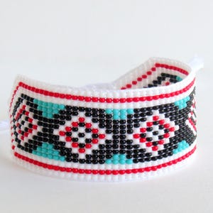 Op de afbeelding: Een witte kralenarmband met een geometrisch patroon. De armband heeft rode, zwarte en turquoise kralen. De armband is ongeveer 3,8 cm breed.