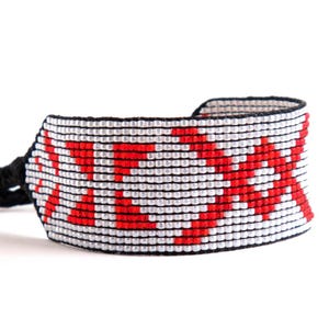 Op de afbeelding: Een brede armband van rode, witte en zwarte kralen. De armband heeft een herhalend patroon van rode kruisen op een witte achtergrond. De randen zijn zwart en de armband heeft een zwarte koordsluiting.