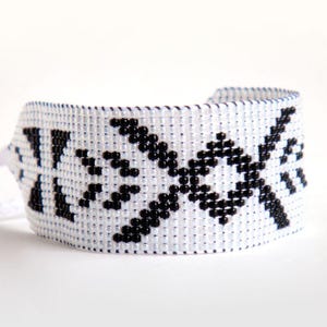 Op de afbeelding: Witte kralen manchetarmband met een geometrisch zwart ontwerp. De armband is gemaakt van kleine, gelijkmatig verdeelde kralen, waardoor een textuur ontstaat. Het ontwerp is symmetrisch en lijkt handgemaakt.