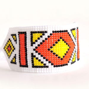Op de afbeelding: Witte kralenarmband met een geometrisch patroon in geel, oranje en zwart. De armband heeft een herhalend ruitontwerp, met een witte achtergrond. De armband is een brede manchetstijl.