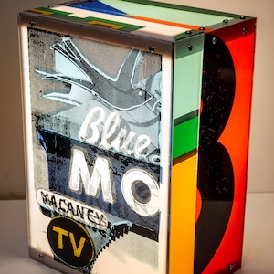 May include: A rectangular, illuminated art piece with a retro motel sign design. The sign reads "Blue MO" and "Vacancy TV" in white and yellow against a blue and black background. The sides are a mix of colours including red, orange, green, and black.