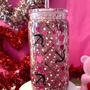 May include: A clear, glitter-filled tumbler with a lid and straw. The tumbler is decorated with pink hearts, white dots, and black bow and arrow designs. The tumbler sits on a white surface against a pink backdrop, with Valentine's Day decorations.