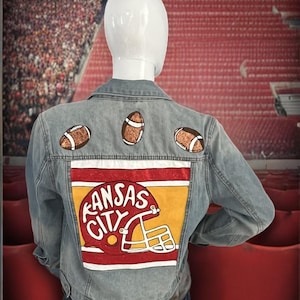 May include: A light blue denim jacket with a painted design on the back. The design features the words "Kansas City" in red and yellow lettering, a red football helmet, and three brown footballs.