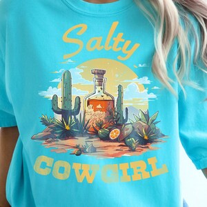 K&ouml;nnte beinhalten: Ein lagunenblaues T-Shirt mit einem W&uuml;sten-Grafikdesign, das Kakteen, eine Flasche und den gelben Schriftzug "Salty Cowgirl" zeigt. Das T-Shirt hat einen Rundhalsausschnitt und kurze &Auml;rmel.