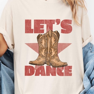 Peut inclure: T-shirt ivoire avec l'inscription rouge "LET'S DANCE" et un motif de bottes de cowboy marron. Le t-shirt a une coupe décontractée et des manches courtes.